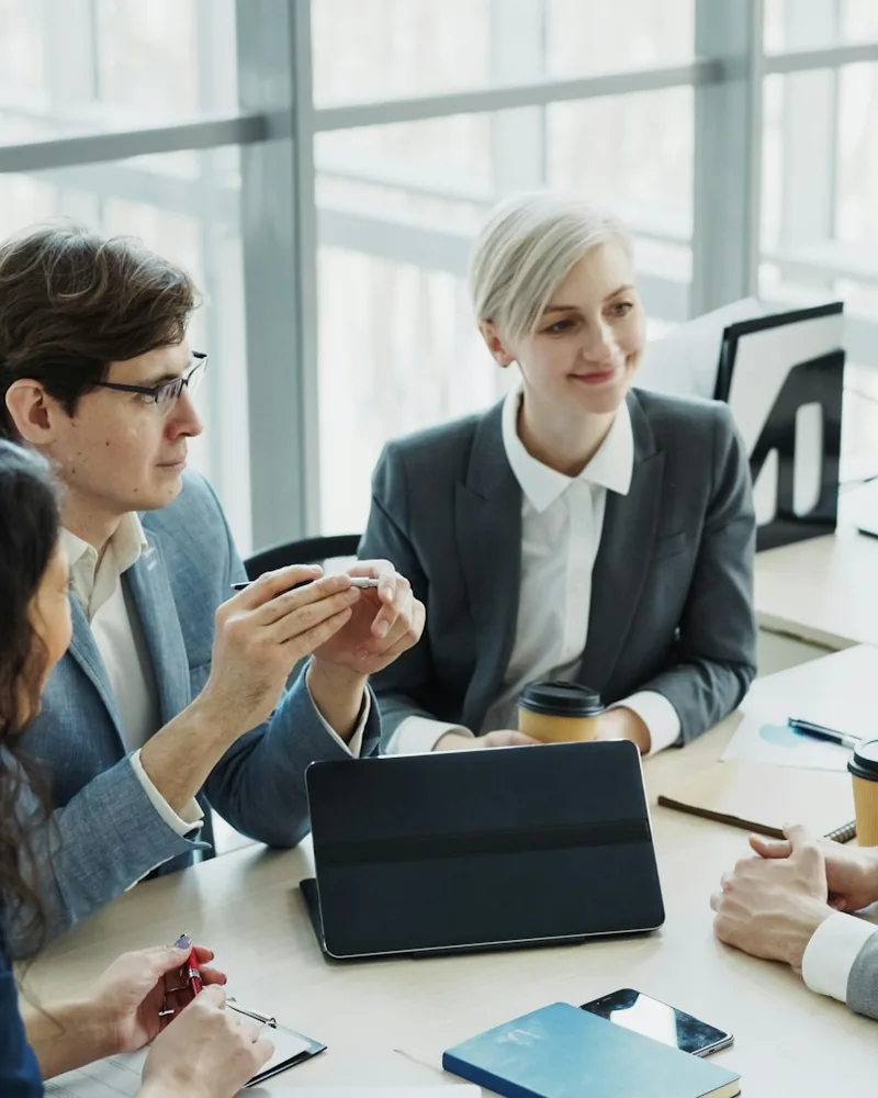 Advisers in business dress talking across a meeting table, with a tablet, printed figures and notepads in front of them.