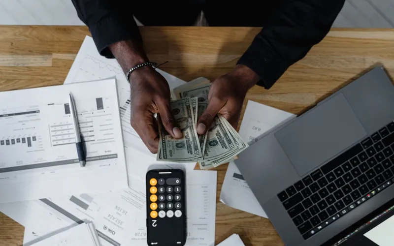 Payroll paperwork, a calculator and a laptop on a desk, seen from above as banknotes are counted.