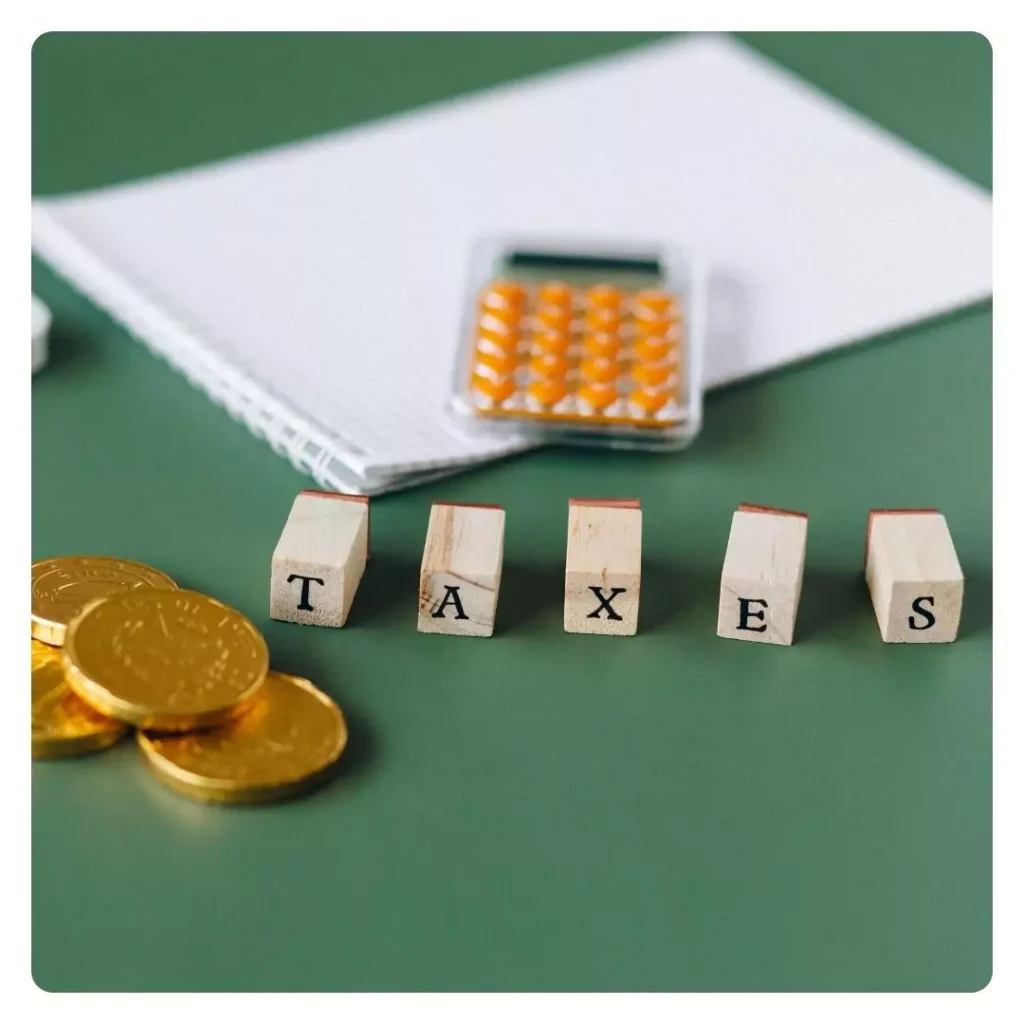 wooden blocks spelling TAXES, accompanied by gold coins, a calculator, and a notebook on a green surface