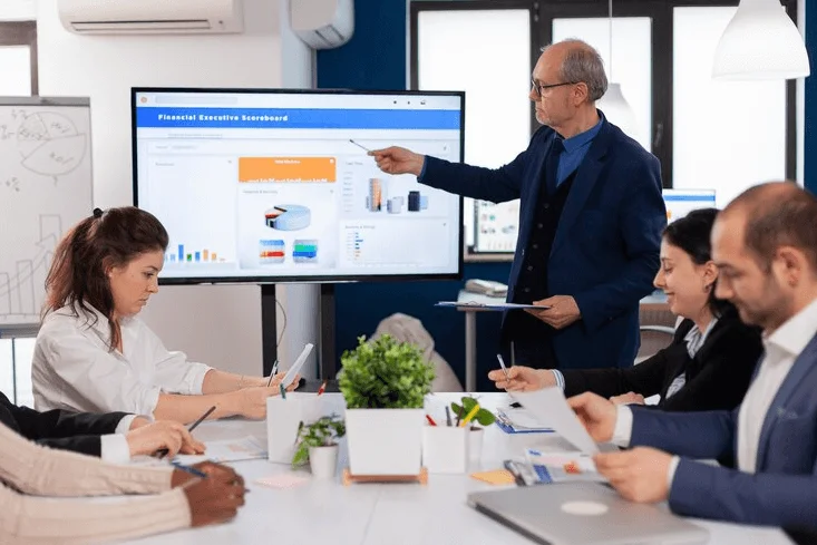 Meeting room scene: man pointing at screen, surrounded by attentive group of people.